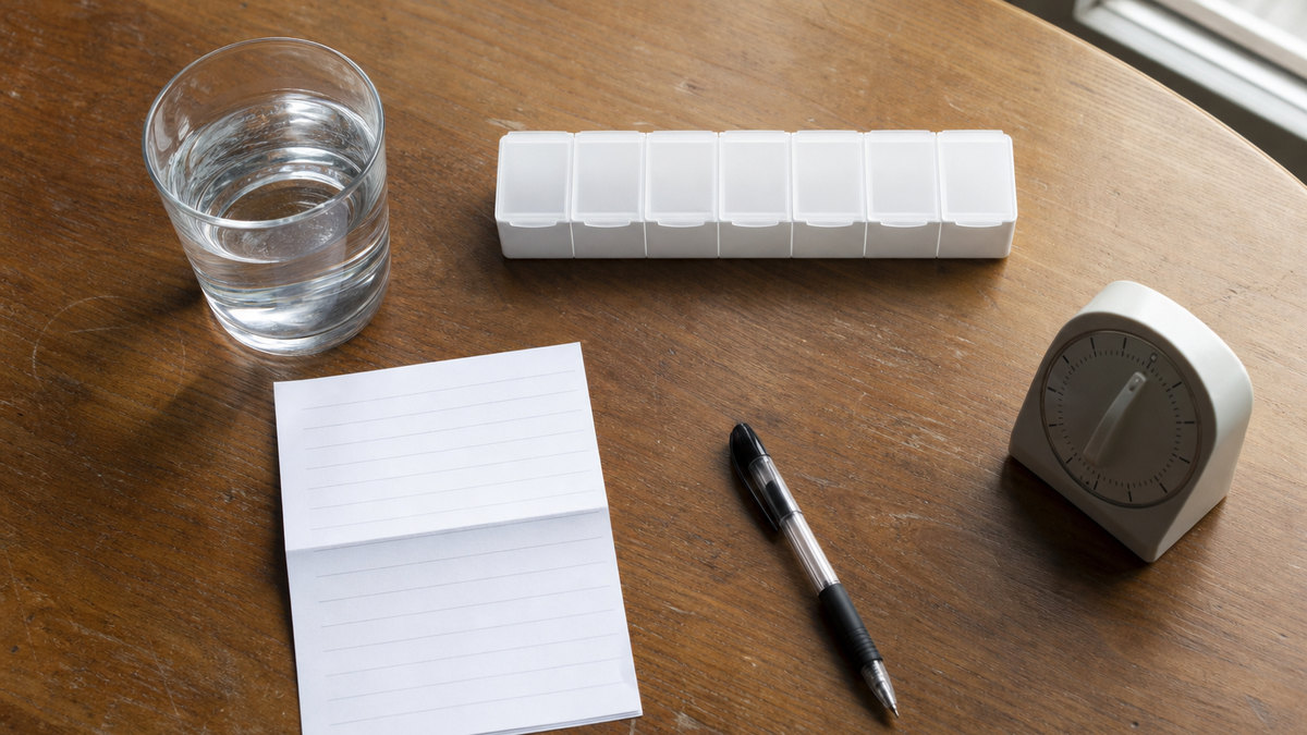 Editorial still life of a weekly pill organizer, a water glass, and unbranded medication-review materials